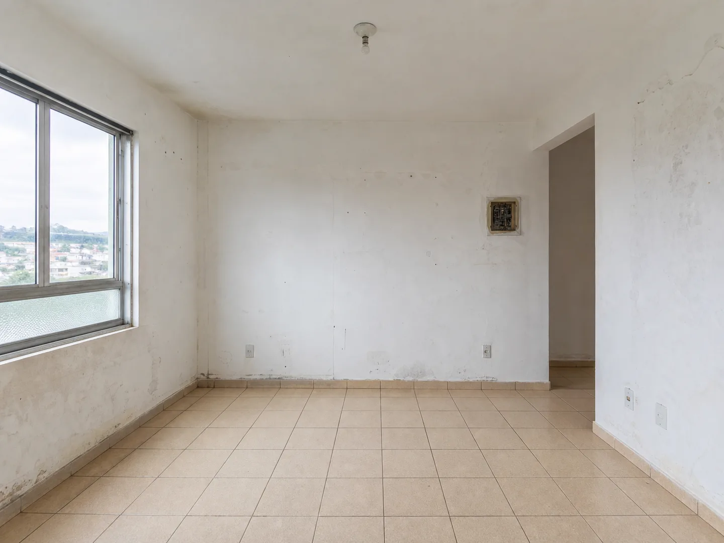 Sala vazia antes da reforma, com piso cerâmico bege antigo, paredes manchadas e quadro de energia aparente.